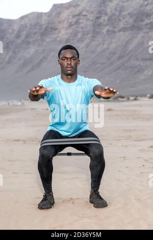 Full body serious African American sportsman squatting with rubber band during fitness workout on Famara Beach in Lanzarote, Spain Stock Photo