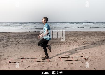 Side view of focused African American sportsman doing ladder drill near sea during fitness training on Famara Beach in Lanzarote, Spain Stock Photo