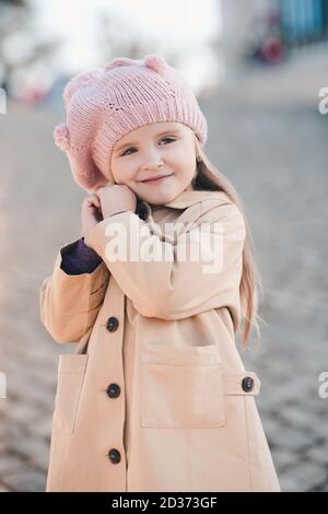Smiling cute baby girl 3-4 year old wearing knitted hat and winter beige coat posing in park outdoors close up. Childhood. Happiness. Autumn season. Stock Photo