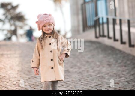 Stylish blonde kid girl 3-4 year old wearing casual clothes: knitted hat and beige winter coat walking in park outdoors close up. Childhood. Autumn se Stock Photo