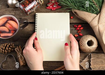 A woman's hands open a notebook against a background of pine branches, cups of mulled wine, cinnamon and berries on a wooden background. Stock Photo