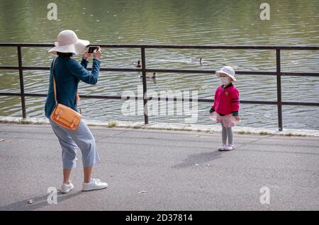 A mother takes cell phone photos of her very cute young daughter. In Kissena Park, Flushing, Queens, New York City.wears, wearing Stock Photo