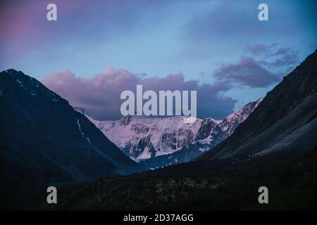 Amazing landscape of sunset with pink snowy mountains and lilac clouds. Atmospheric highland scenery with great mountain silhouettes under purple dawn Stock Photo