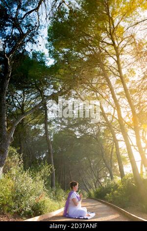 Woman meditating sitting in the lotus pose with her hands in front of her chest, in anjali mudra, in a forest path at sunset Stock Photo