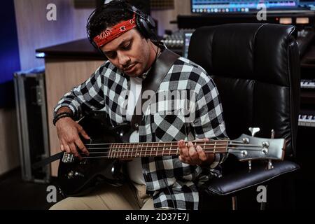 Portrait of focused young man, male artist in headphones playing guitar while sitting in recording studio Stock Photo