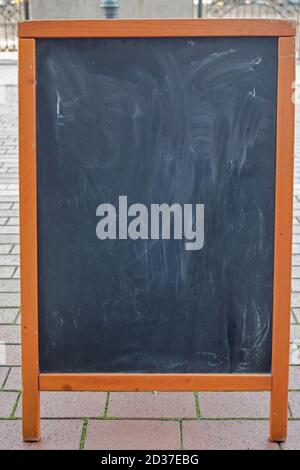 Black chalkboard with traces of chalk in brown wooden frame standing on street of beige tiles in blurred background of fence. Mockup for menu design. Stock Photo