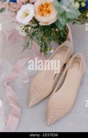 Close-up of the bride's ballet flats with shiny stones with a bouquet of flowers and a silk ribbon on a white background. Stock Photo