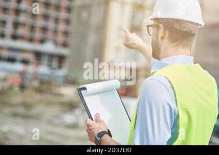 Building industry. Rear view of a young civil engineer or construction supervisor wearing helmet pointing at construction site Stock Photo
