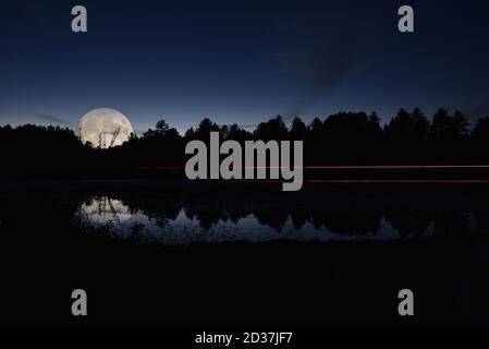 Reflection of light trails from a vehicle on a small lake at night, set against trees and night sky. Stock Photo