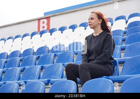 A young woman in a black tracksuit with long hair sits on the stadium stand alone and watches a sports game Stock Photo