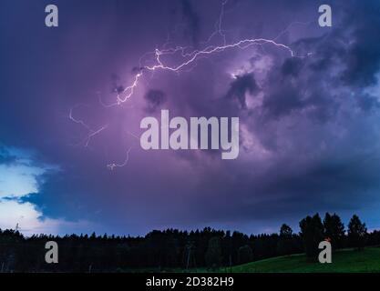 A powerful lightning bolt striking down from dark purple storm clouds over a silhouette of a distant forest. Stock Photo