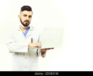 Man with concentrated face in white coat. Doctor with beard type on his white laptop. Treatment and medical technologies concept. Physician in surgical mask isolated on white background, copy space Stock Photo