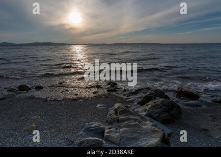 A sandy beach with some large rocks scattered throughout. The water has some small waves. The sky is setting and the sun is going down. Stock Photo