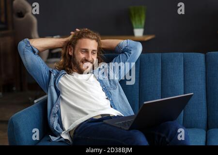 Time to relax. A curly-haired guy in a casual style, sitting on the couch at home, smiling and relaxing. Pleasant leisure time. Rest at home Stock Photo