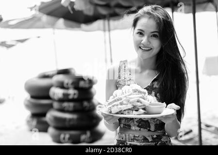 Young happy beautiful Asian woman smiling while holding plate of steamed freshly cooked crabs at the beach Stock Photo