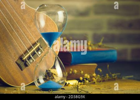 A vintage hourglass and ukulele with an old book and brass pen on a wooden table and brick background in the morning. Closeup and copy space. The conc Stock Photo