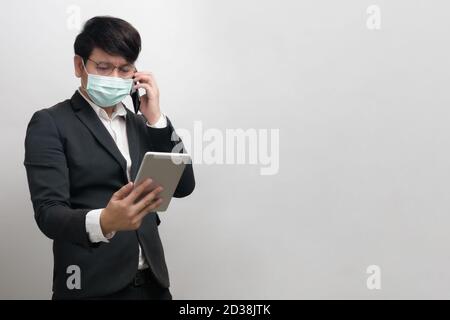 Asian businessman wearing surgical face mask in formal black suit jacket, using smart phone to talking by right hand and look at the tablet, studio li Stock Photo