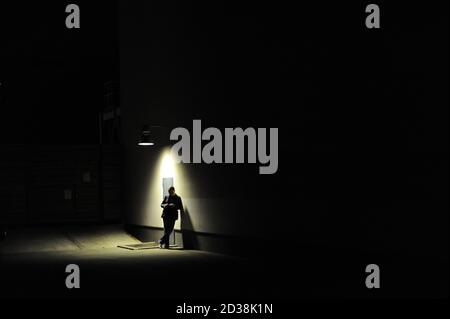 Man leaning against a wall, standing under a street lamp at night, silhouetted by the light Stock Photo