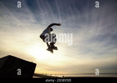 Unrecognizable roller on the extreme roller skates performing acrobatic jump at sunny sky - Guy at extreme sport competition on sunny afternoon - Alternative lifestyle concept on warm sunshine colors. Warm toned image. Stock Photo