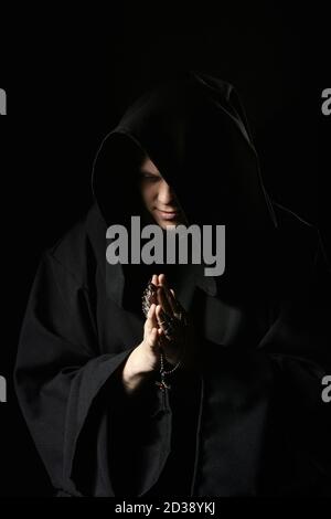 Assassin in dark clothes in church. Sorcerer in black mantle doing mysterious rite in dark room. Monk prays in darkness. Magic ritual in shadow. Sinne Stock Photo