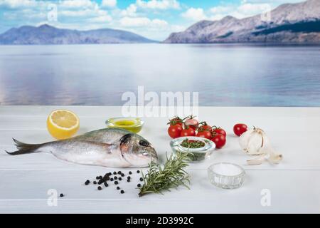 a sea bream and with products for the preparation of a recipe on a white wooden table with the sea bay in the background Stock Photo