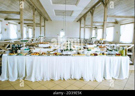 Large wedding area in pastel colors with a variety of colored sweets on a table covered with a white tablecloth in a bright room with wooden beams. Ca Stock Photo
