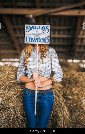 Faceless blonde woman in cowboy clothes holding a wooden board with the inscription dallas cowboys in front of her face on a background of hay. Rustic Stock Photo