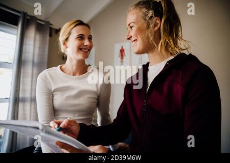 Young female patient and physiotherapist smiling while giving instructions sitting on massage bed in studio. Stock Photo