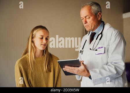 Professional Doctor completing check up and diagnosing patient, typing on digital tablet and discussing results with female patient Stock Photo