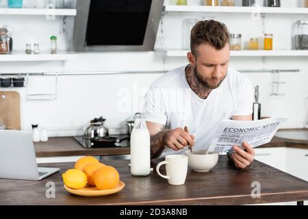 tattooed man in white t-shirt having breakfast in kitchen and reading newspaper Stock Photo