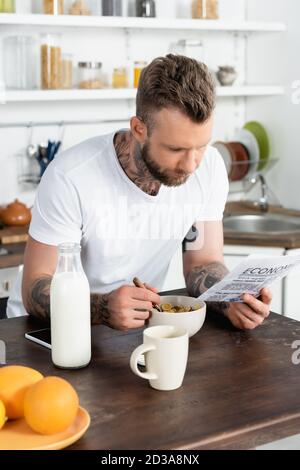selective focus of bearded, tattooed man reading newspaper while having breakfast in kitchen Stock Photo