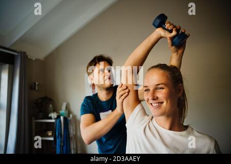 Female patient smiling while correcting arm exercises, assisted my male physiotherapist in pilates studio. Stock Photo