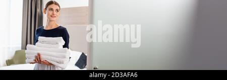 horizontal image of young maid in apron looking at camera and holding clean towels in hotel room Stock Photo