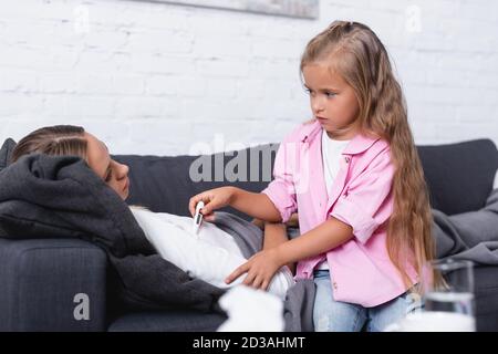 Selective focus of child holding thermometer near mother lying on couch Stock Photo