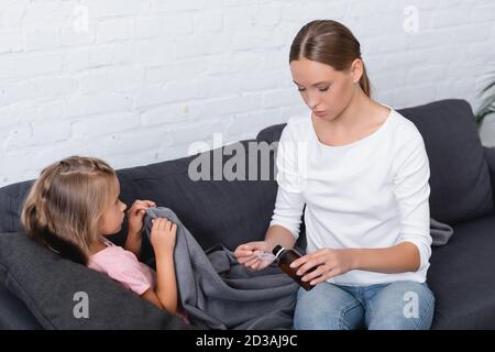 Sick child with blanket lying near mother pouring syrup on couch Stock Photo