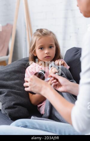 Selective focus of woman pouring syrup near sick daughter with blanket on sofa Stock Photo