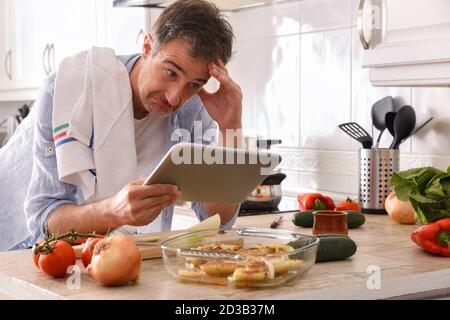Novice chef looking at a tablet with an inept face resting his elbow on the kitchen bench touching his forehead Stock Photo