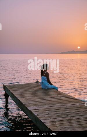 Crete Greece, young romantic couple in love is sitting and hugging on wooden pier at the beach in sunrise time with golden sky. Vacation and travel Stock Photo