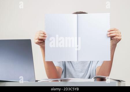 Kid hands holding open notebook with blank white pages Boy sitting at table with books and laptop computer Stock Photo