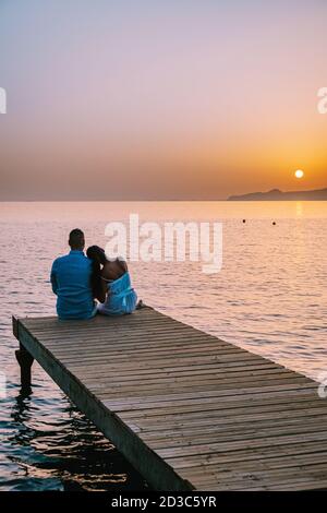 Crete Greece, young romantic couple in love is sitting and hugging on wooden pier at the beach in sunrise time with golden sky. Vacation and travel Stock Photo