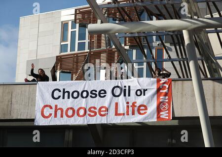 Three members of Extinction Rebellion Scotland who have climbed on to Holyrood, the Scottish Parliament in Edinburgh, and draped a banner reading 'Choose Oil or Choose Life' from an overhang above the public entrance in a protest against fossil fuels. Stock Photo