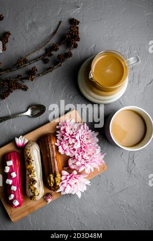 Top view of delicious eclairs on wooden kitchen board decorated with chrysanthemums, cup of coffee and branches of flowers on gray textured background Stock Photo