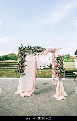 Elegant wedding arch made of pink flowers of hydrangea, roses and greenery on a spacious green lawn in the park on the background of the lake. Beautif Stock Photo