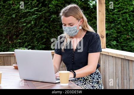 Young blond woman wearing blue face mask, sitting at table, using laptop computer. Stock Photo