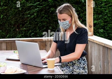 Young blond woman wearing blue face mask, sitting at table, using laptop computer. Stock Photo