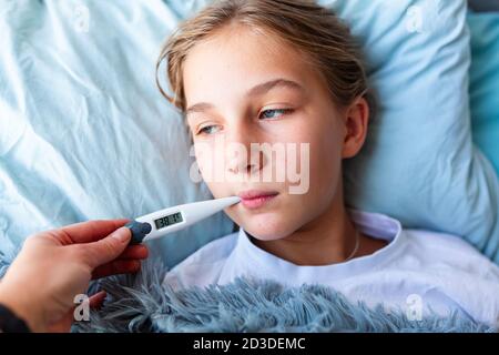 Sick teenage little girl with high fever and headache laying in bed and holding thermometer in her mouth. Stay at home during corona virus epidemic if Stock Photo