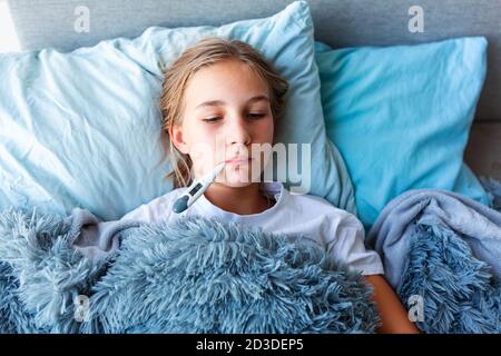 Sick teenage little girl with high fever and headache laying in bed and holding thermometer in her mouth. Stay at home during corona virus epidemic if Stock Photo