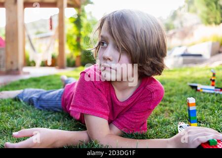 Boy with brown hair lying on lawn, playing with building blocks. Stock Photo