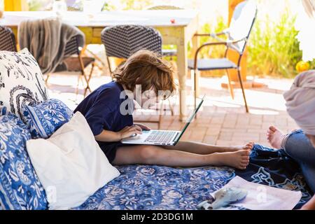 Boy with brown hair sitting on outdoor bed, doing homework on laptop. Stock Photo