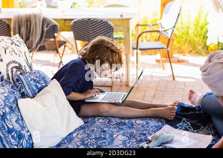 Boy with brown hair sitting on outdoor bed, doing homework on laptop. Stock Photo